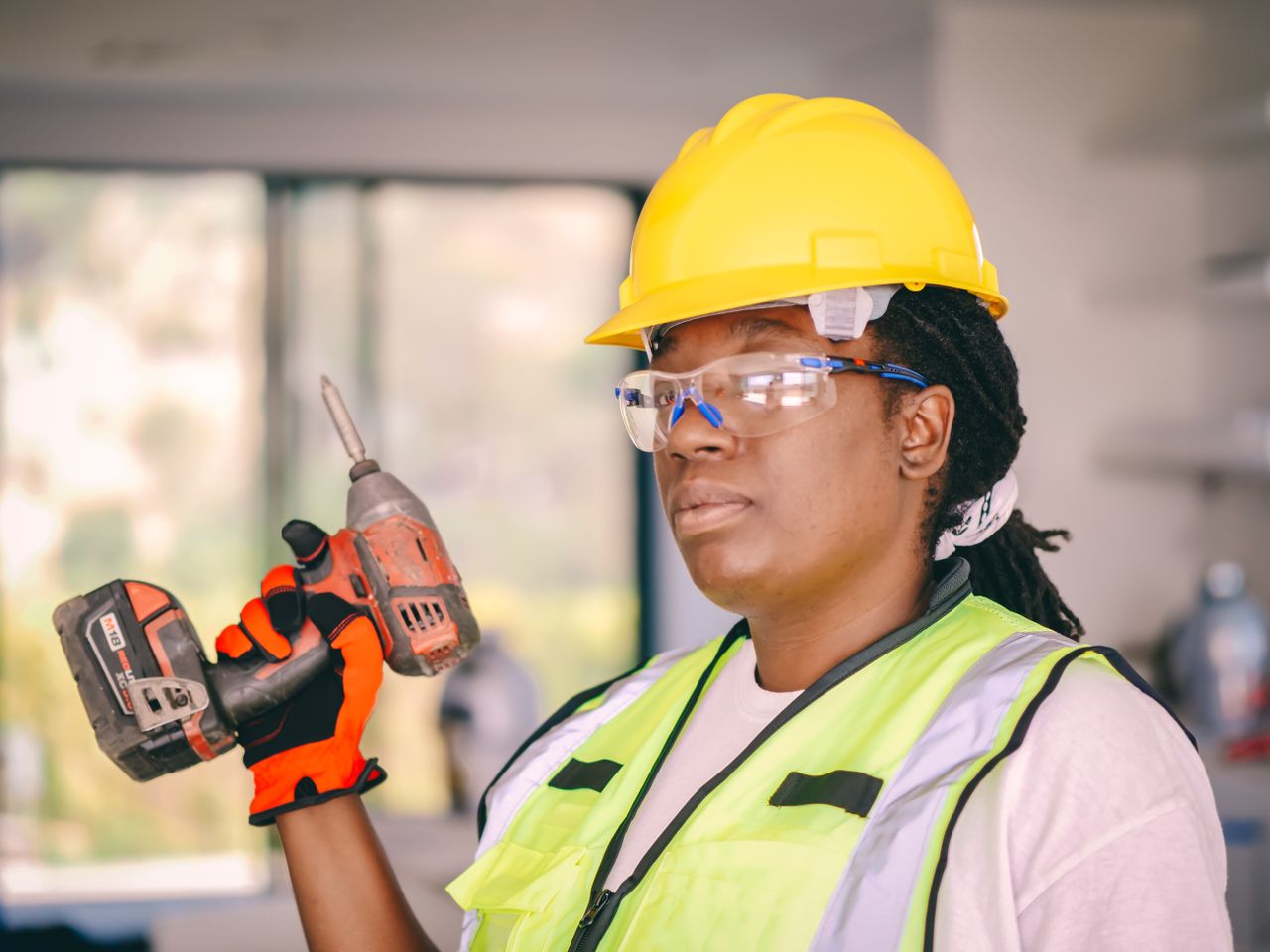 Confident female construction worker wearing safety gear and holding a drill indoors.